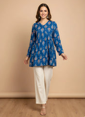 Woman wearing a blue patterned top and white pants against a beige background