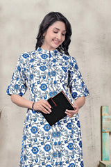 Woman wearing a blue and white floral dress holding a black book against a beige wall.