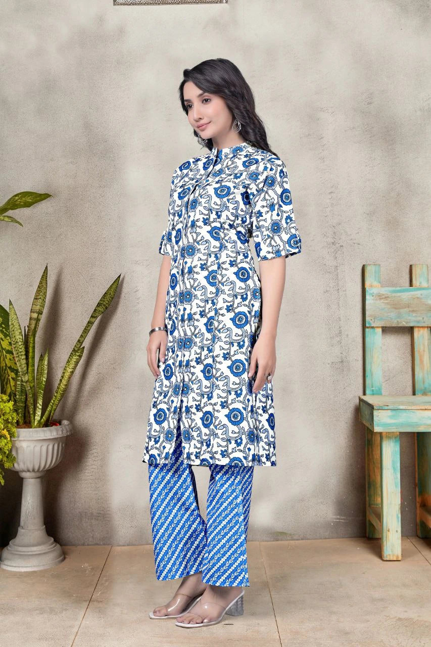 Woman wearing a blue and white floral outfit standing against a gray wall with a plant and wooden chair in the background.
