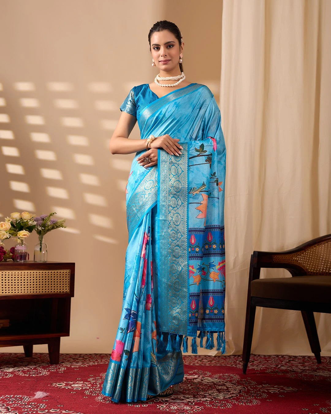Woman wearing a blue saree with a pattern, standing in a room with a beige curtain and wooden furniture.