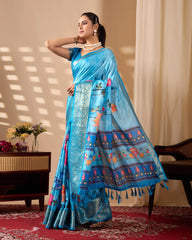 Woman wearing a blue saree with intricate patterns in a room with a table and flowers.