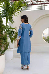 Woman in a blue traditional outfit standing on a rooftop with plants and architectural elements.
