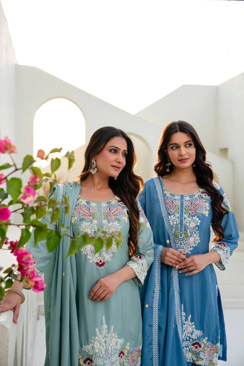 Two women in traditional embroidered dresses standing outdoors with a white architectural background.
