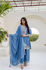 Woman in a blue traditional outfit with floral patterns standing in a bright, minimalistic setting.