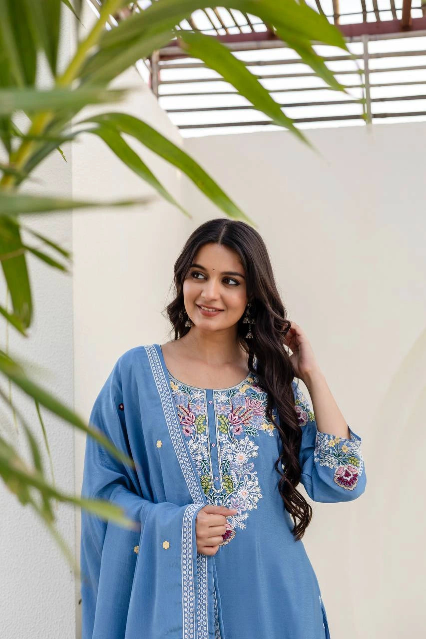 Woman in a blue embroidered outfit standing outdoors with greenery.