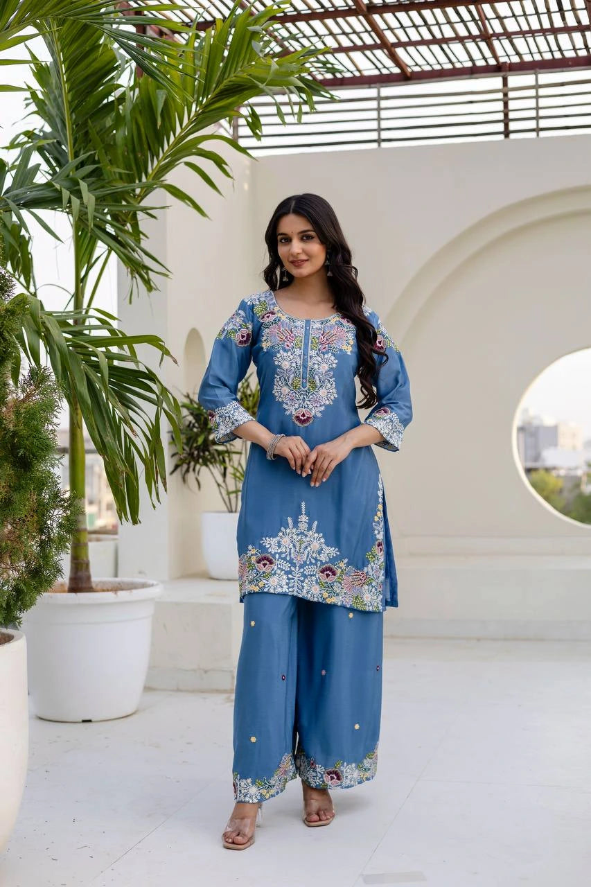 Woman in a blue embroidered outfit standing in an outdoor setting with plants and a white wall.