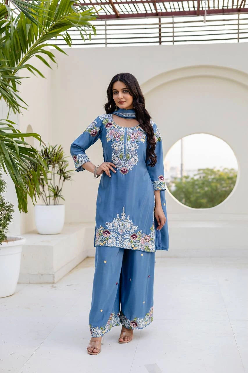 Woman in a blue embroidered outfit standing in a bright, minimalistic outdoor setting with plants.