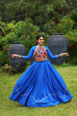 Woman in a blue traditional outfit with colorful embellishments standing outdoors.