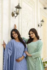 Two women in blue and green traditional outfits standing against a white wall.