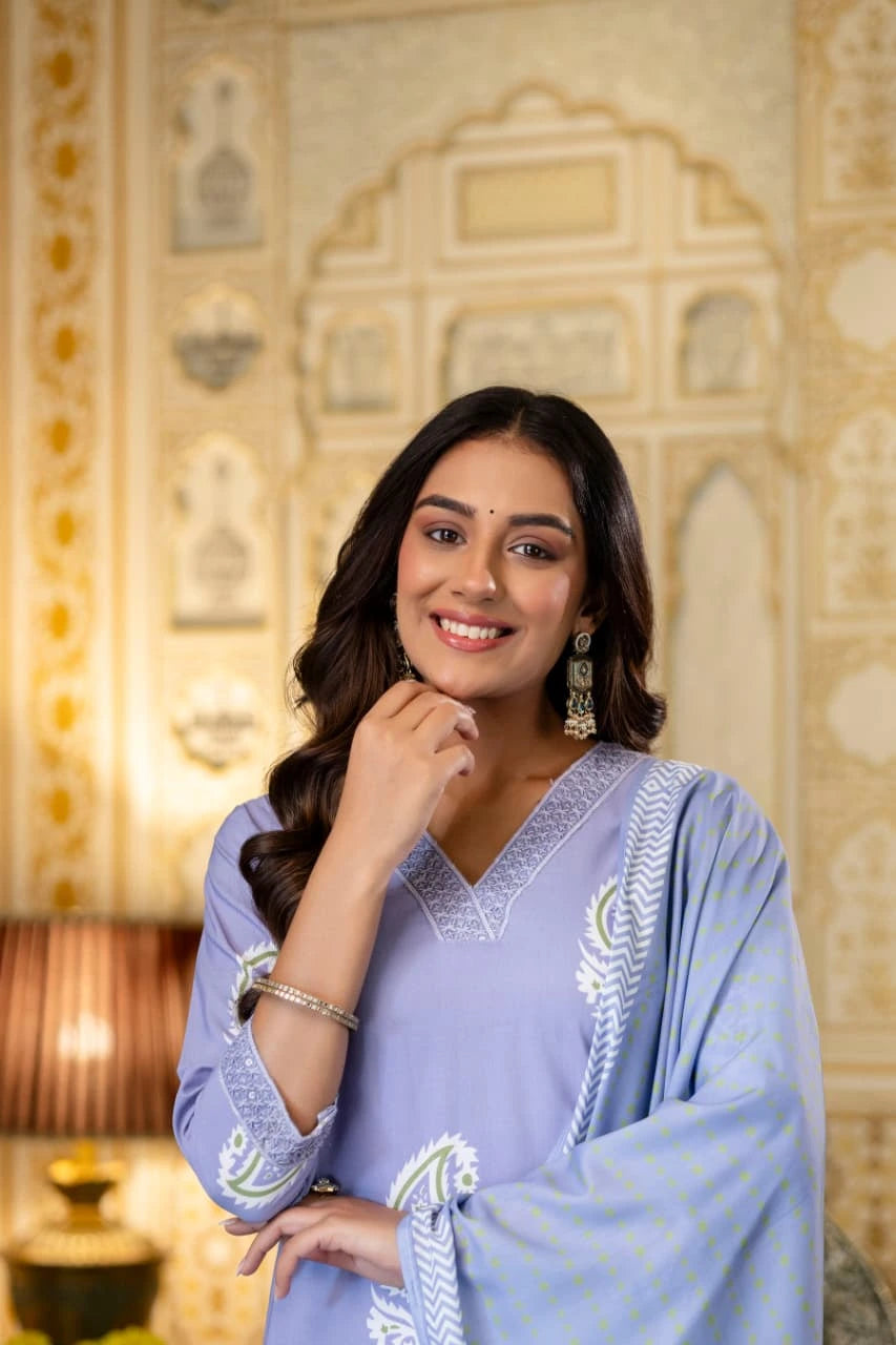 Woman in a light blue traditional outfit with intricate patterns, standing in front of an ornate wall.