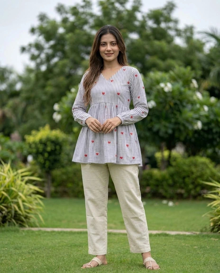 Woman standing outdoors in a garden wearing a striped shirt and beige pants.