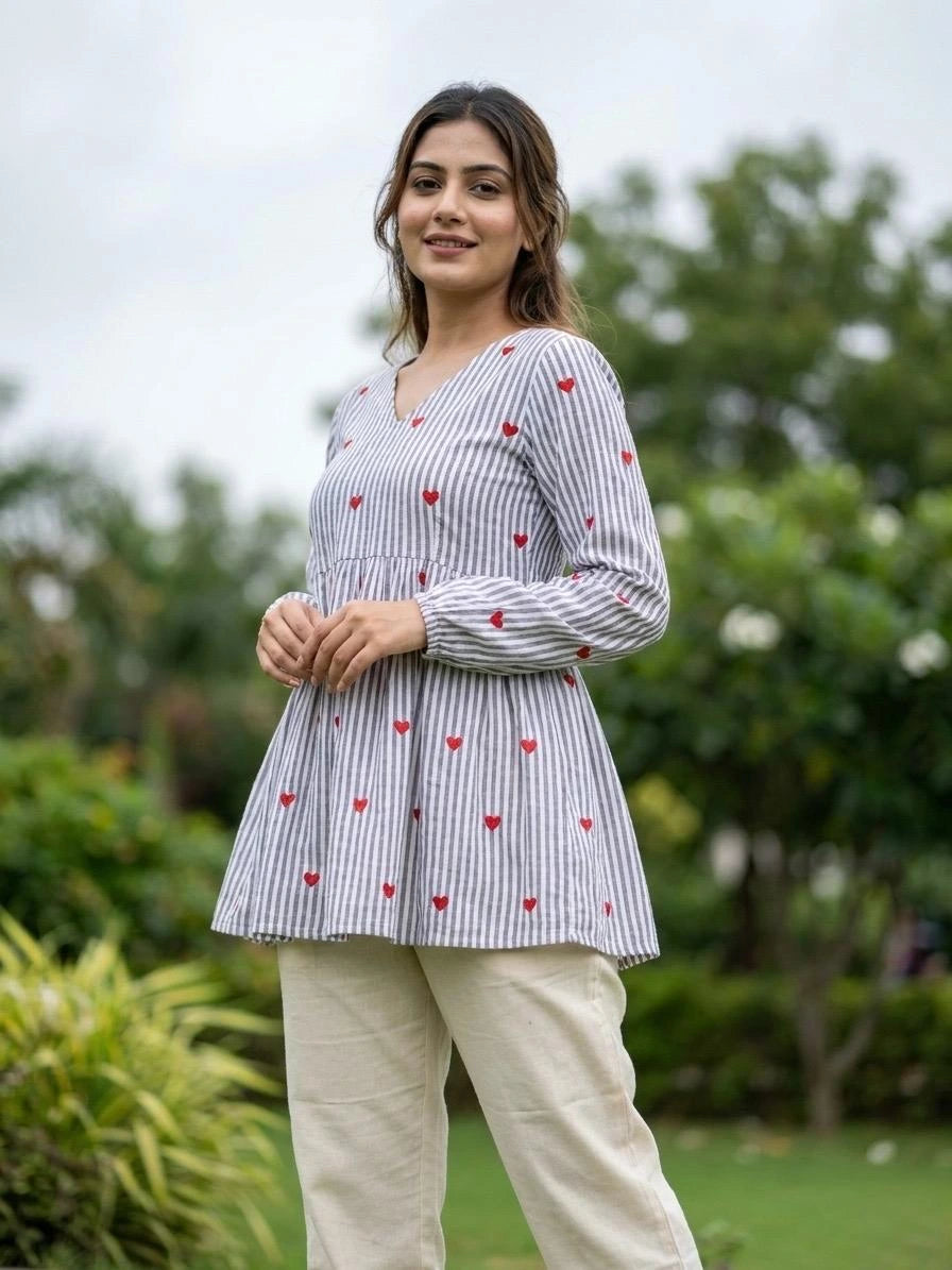 Woman wearing a white blouse with red heart patterns and beige pants standing outdoors.
