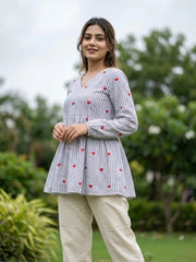 Woman wearing a white blouse with red heart patterns and beige pants standing outdoors.