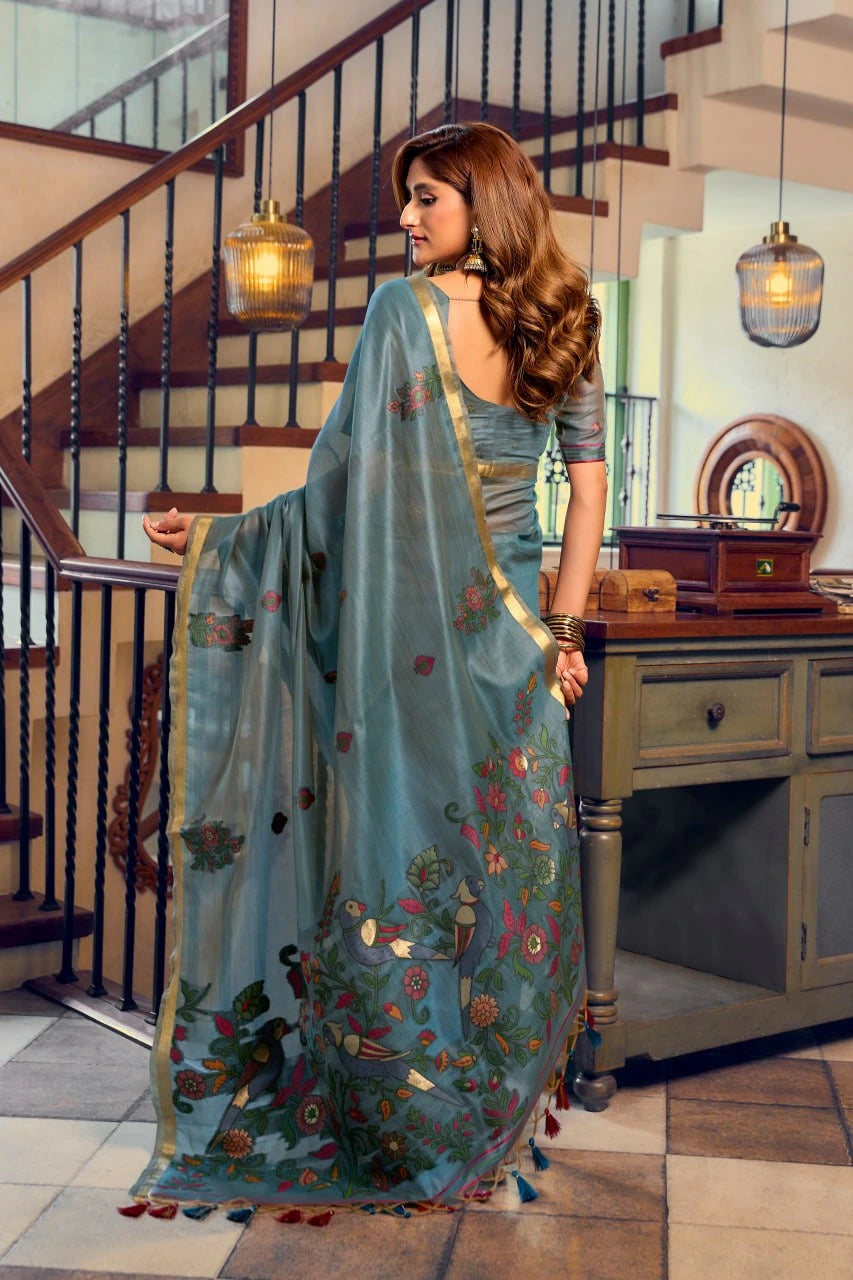 Woman in a blue saree with floral patterns standing in a room with wooden staircase and furniture.