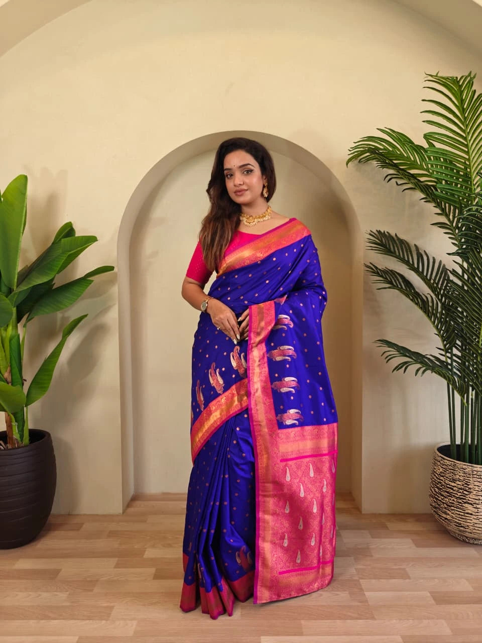 Woman in a blue and pink saree standing in a room with plants.