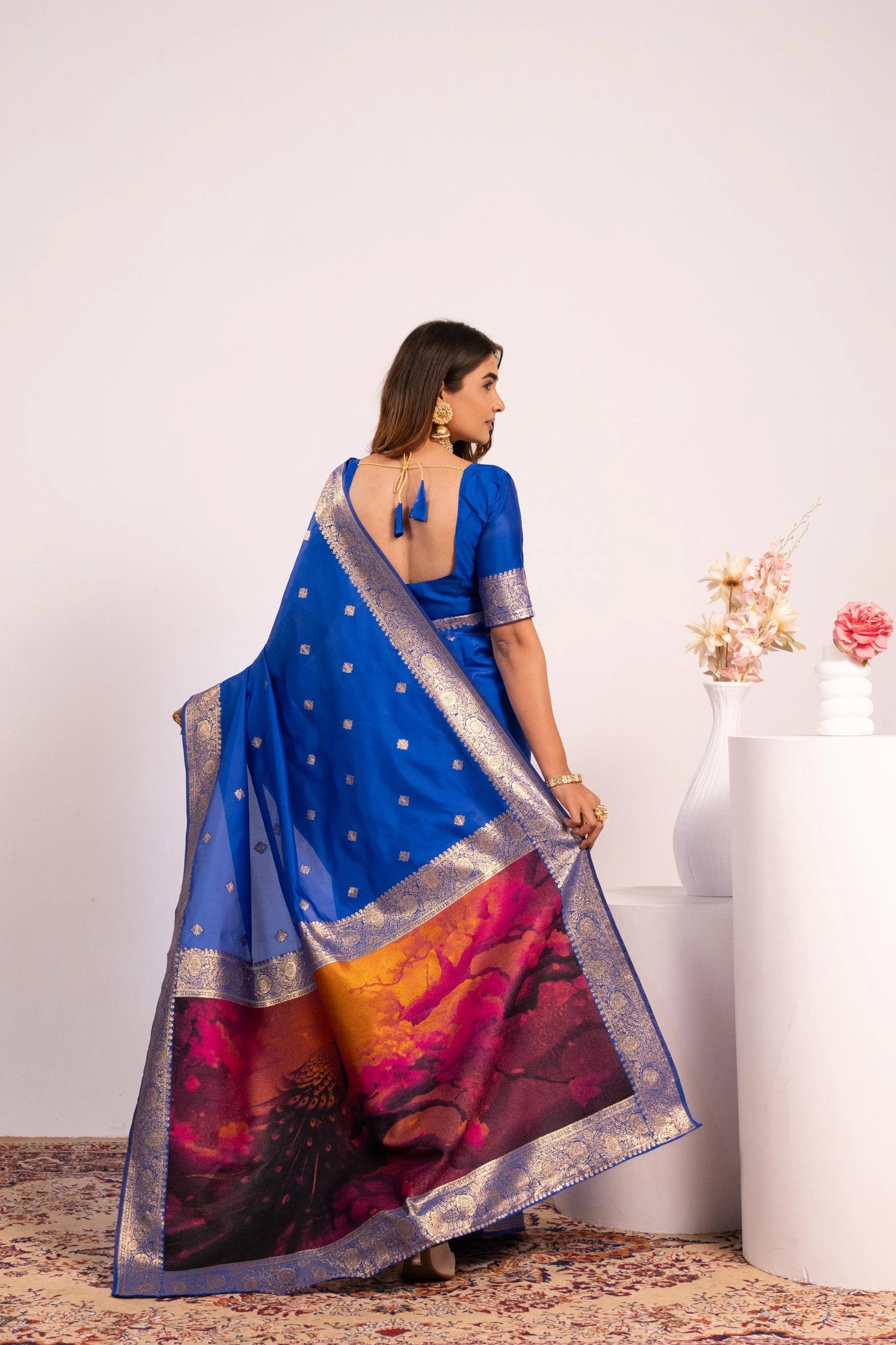 Woman in a blue saree with a colorful pattern, standing against a white wall.