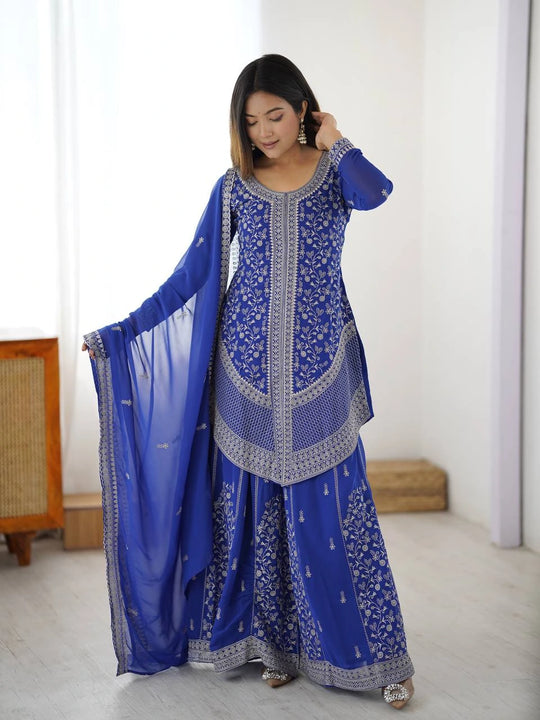 Woman wearing a blue embroidered traditional outfit in a minimalistic room.