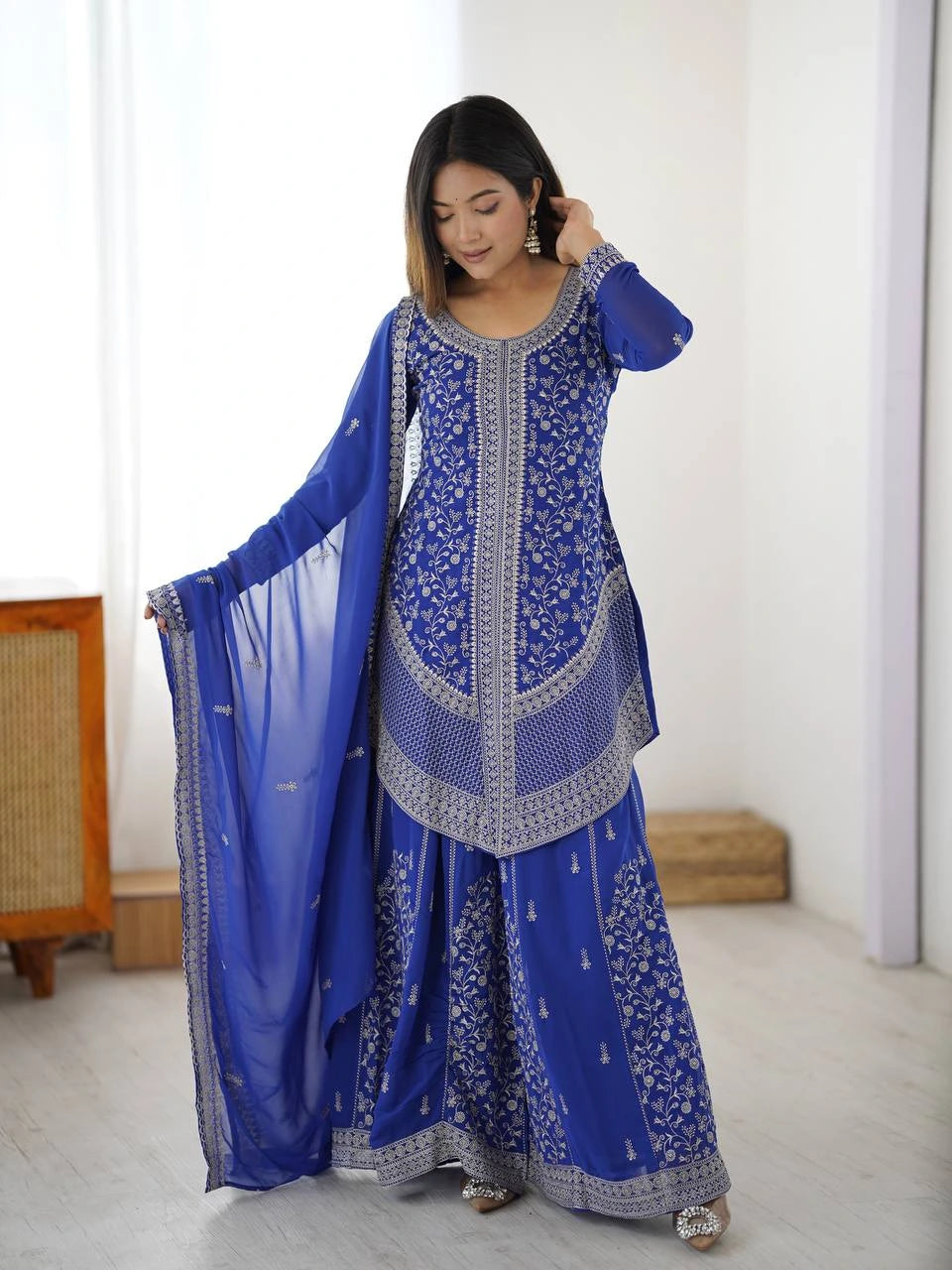 Woman wearing a blue embroidered traditional outfit in a minimalistic room.