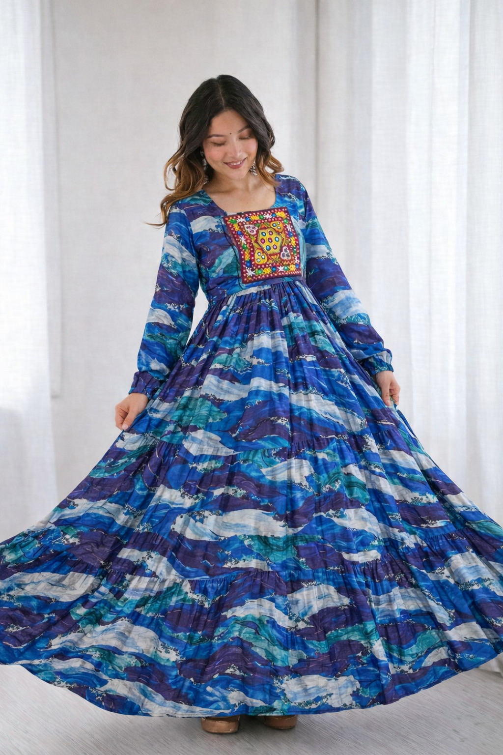 Woman wearing a long, colorful dress with a patterned top against a white curtain background