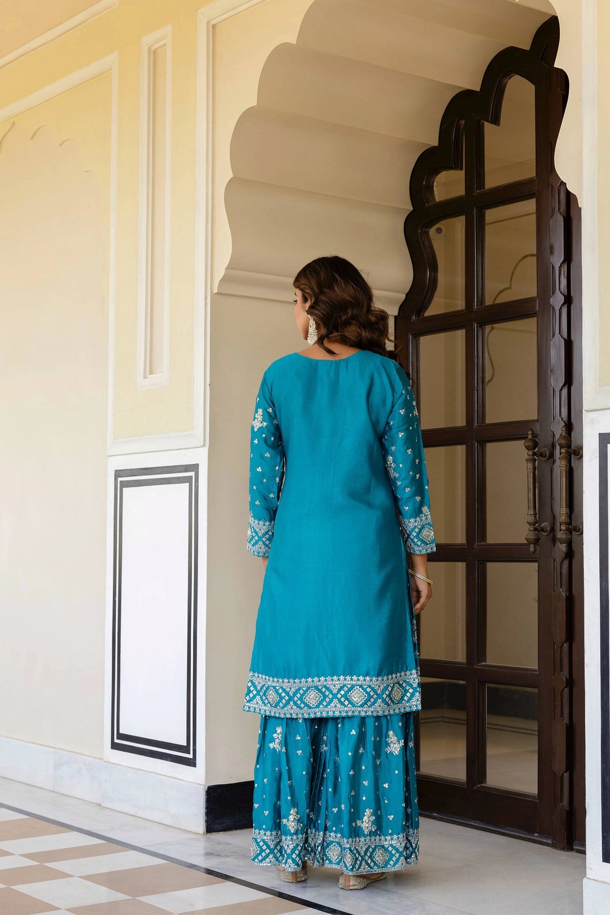 Woman in a blue traditional outfit standing in a decorative interior setting.