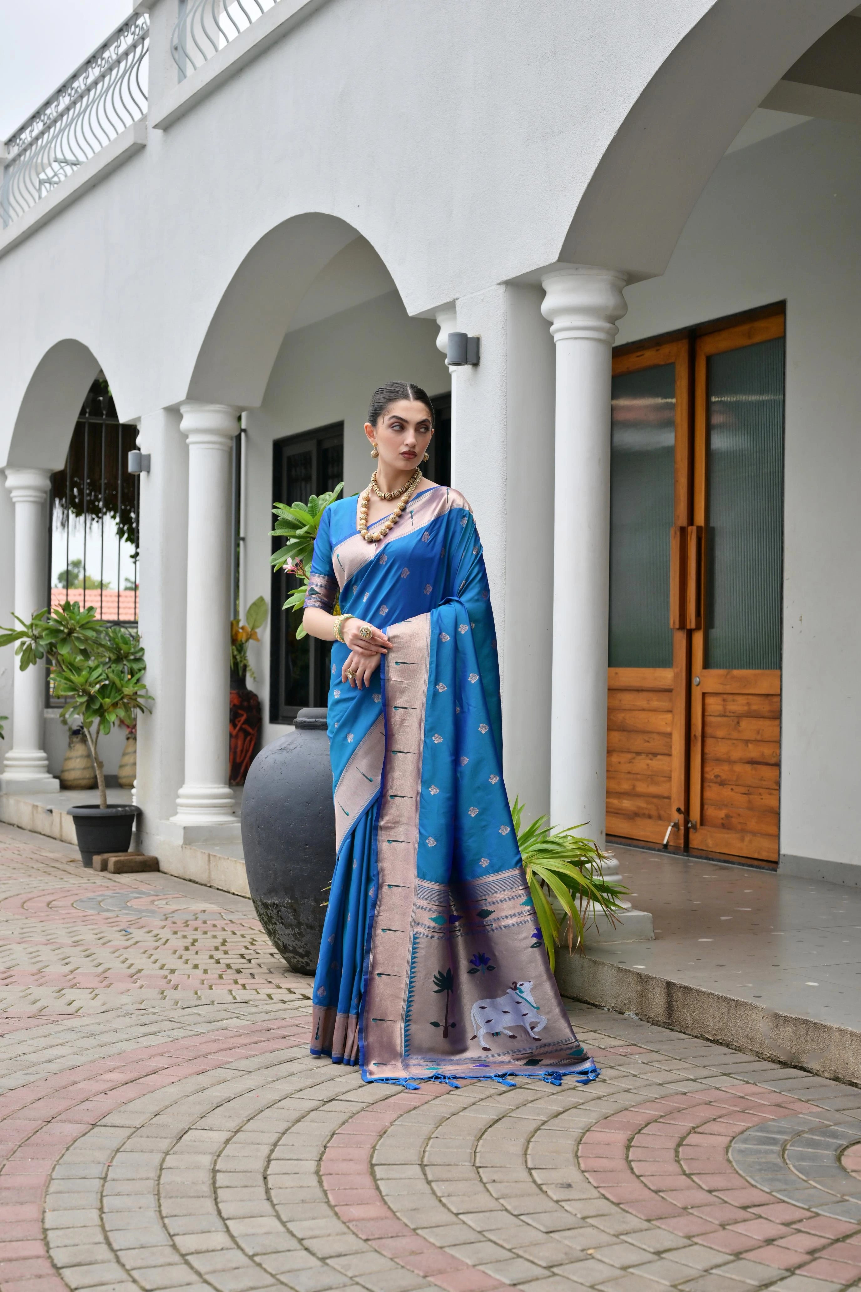 Woman in a blue saree standing in front of a white building with arches and wooden doors.