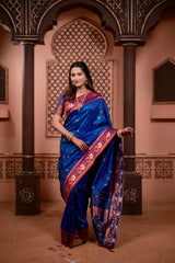 Woman in a blue saree with red border standing in an ornate architectural setting.