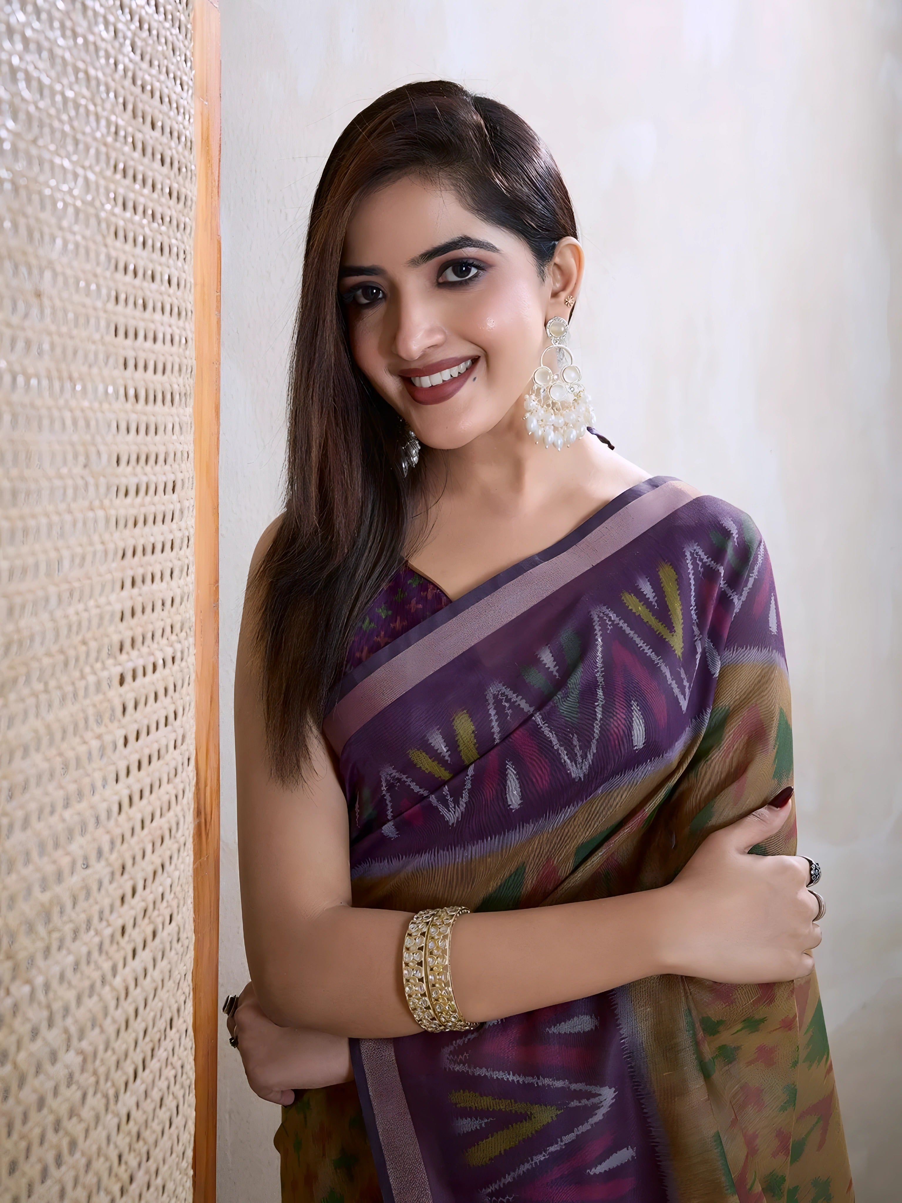 Woman wearing an olive green georgette saree featuring a striking purple geometric border and abstract patterns, paired with a matching blouse, traditional silver earrings, and stacked bangles. She stands elegantly in a contemporary studio setting with a textured woven screen, a neutral-toned wall, and a rustic wooden floor, showcasing graceful draping and a flowing pallu.
