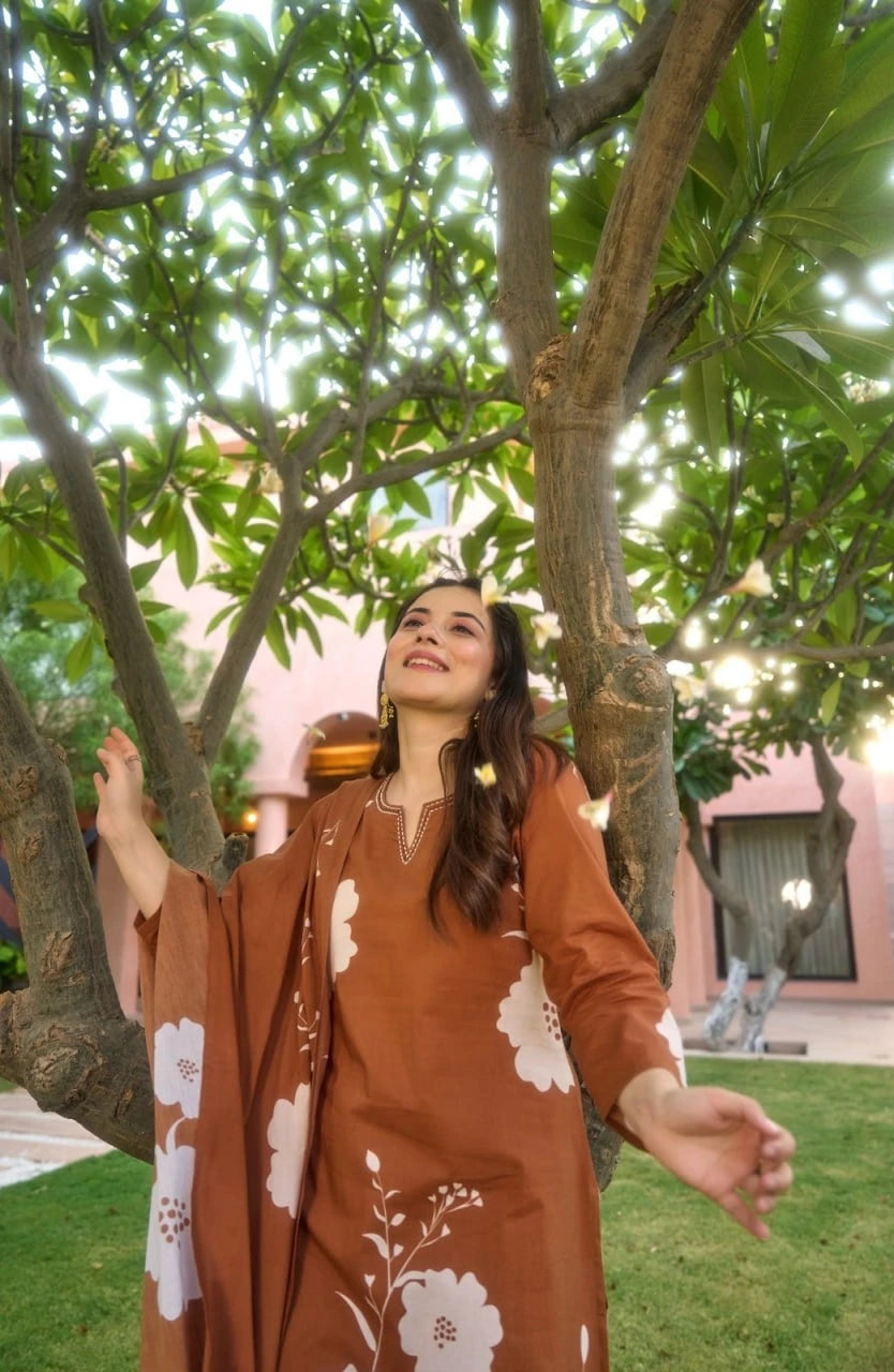 A woman stands beneath a lush green tree, looking upward with a smile as small white flowers fall around her. She is wearing a terracotta-colored kurta featuring large, stylized white floral motifs and a delicate embroidered neckline.