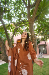 Woman in a brown floral dress standing under a tree with a pink building in the background