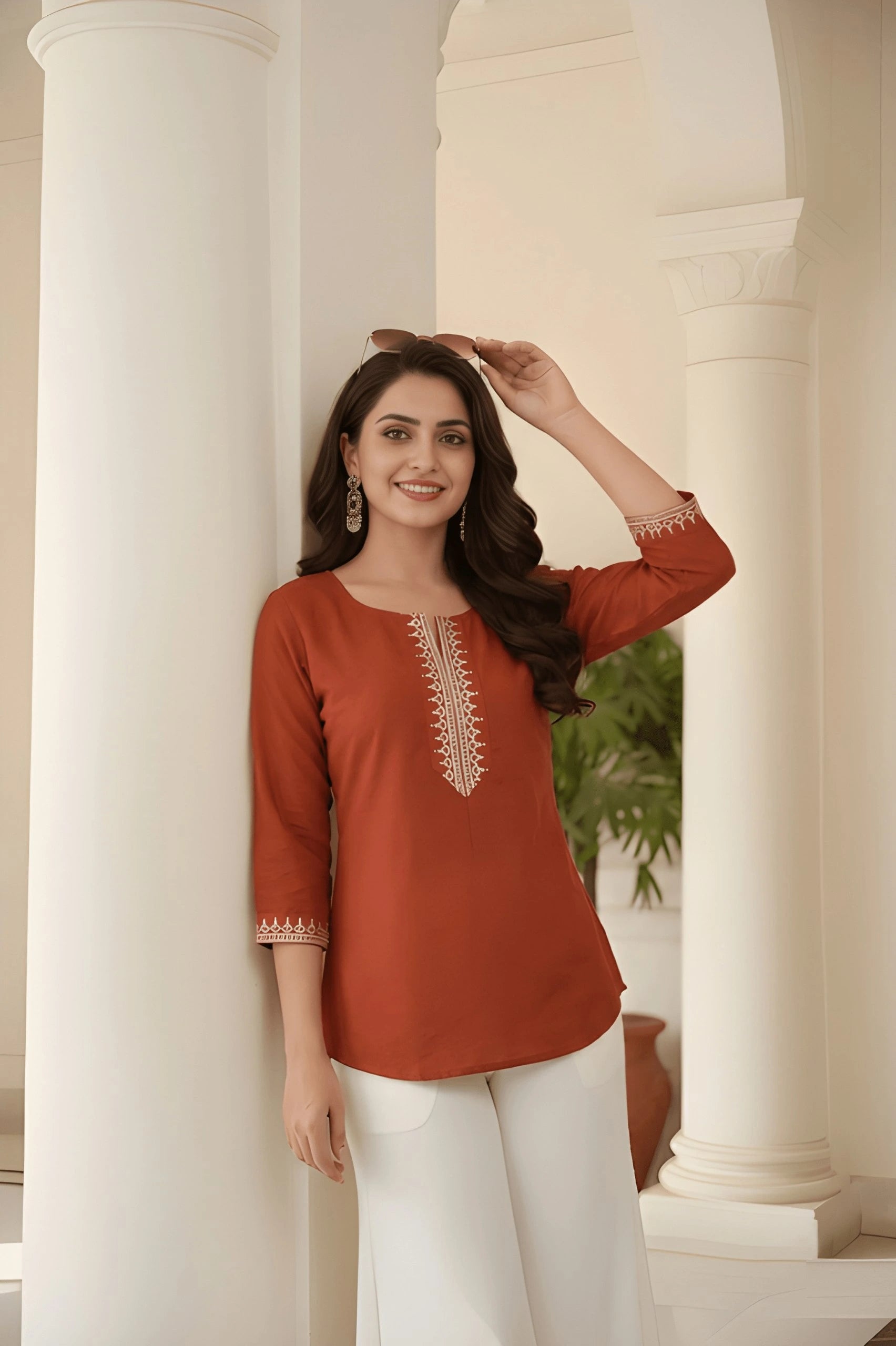 Woman wearing a rust-colored top with white pants standing in a classical architectural setting.