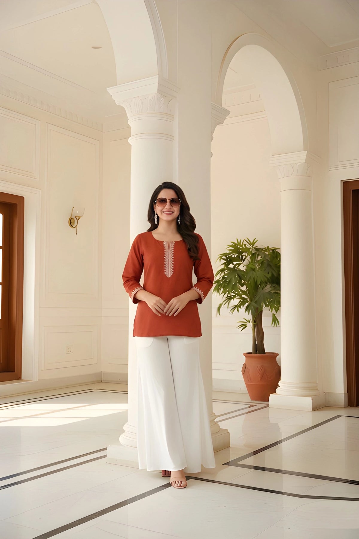 Woman in a red top and white pants standing in a sunlit hallway with columns and a plant.