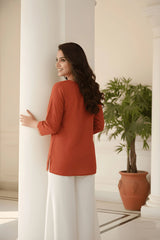 Woman in a red top and white pants standing in a room with a plant and columns.