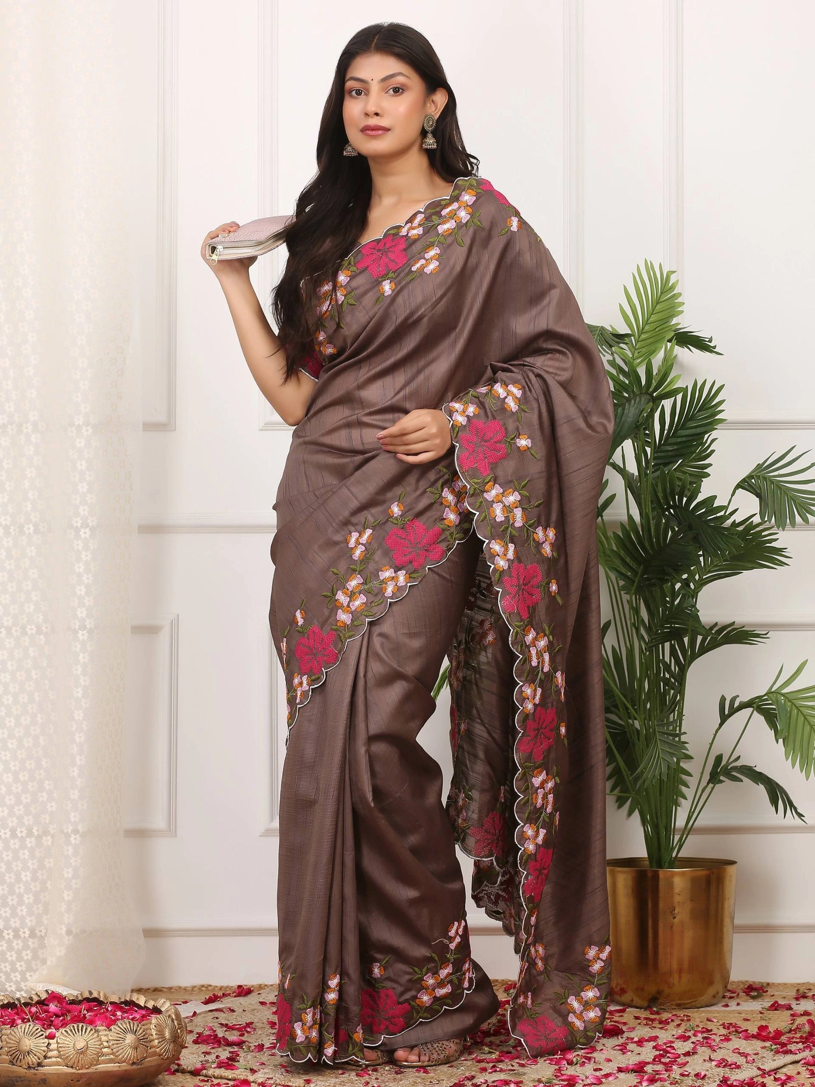Woman wearing a brown saree with floral patterns in a room with a plant and decorative items.