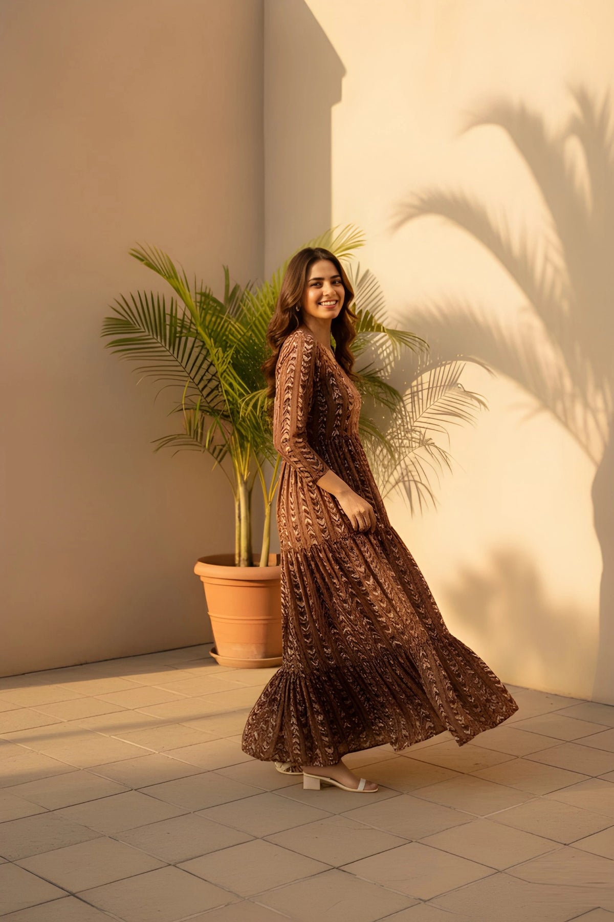 Woman in a long, patterned dress standing in front of a light-colored wall with a potted plant.