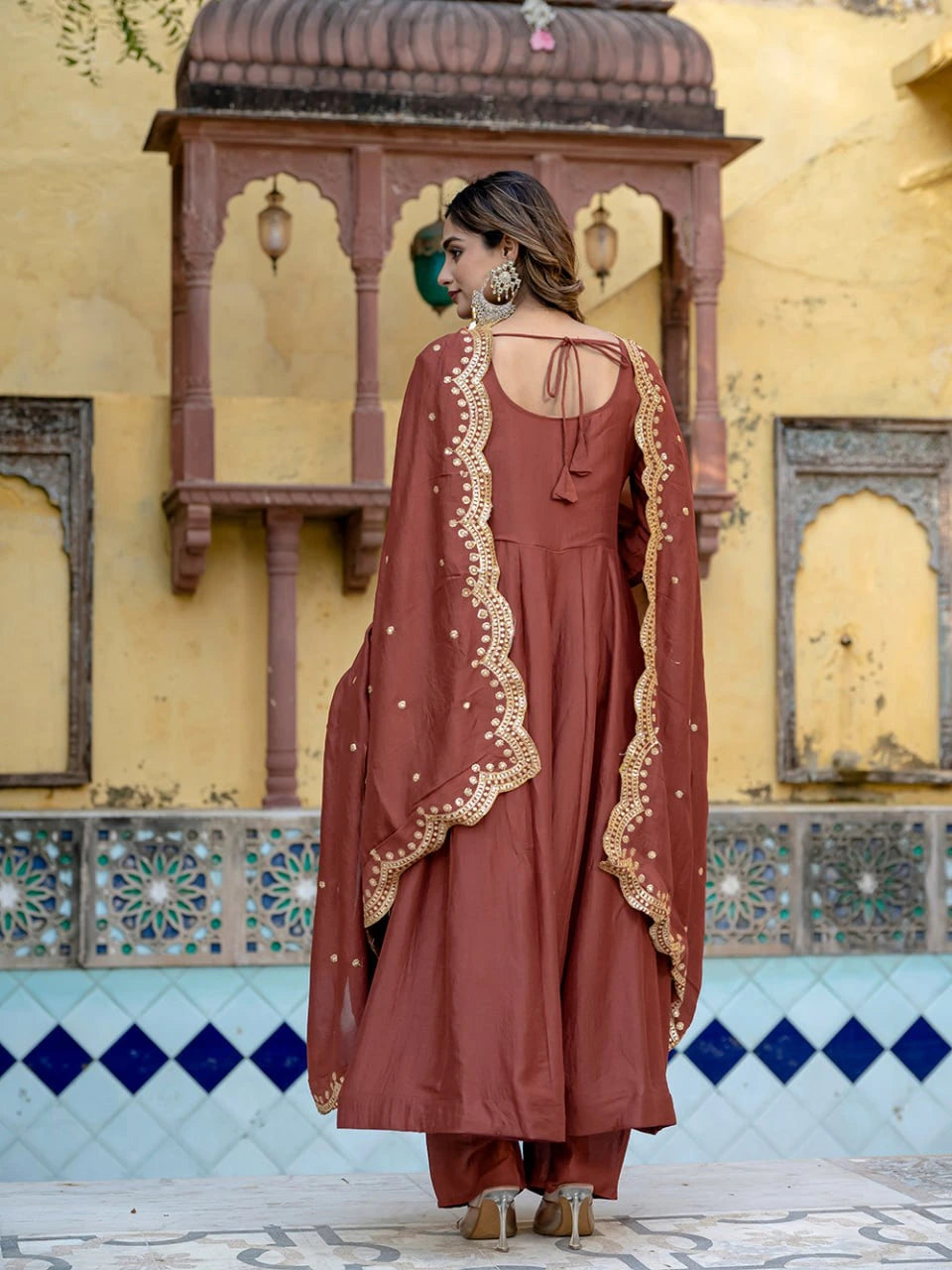 Woman in a rust-colored traditional outfit with gold embroidery standing in front of a decorative wall.