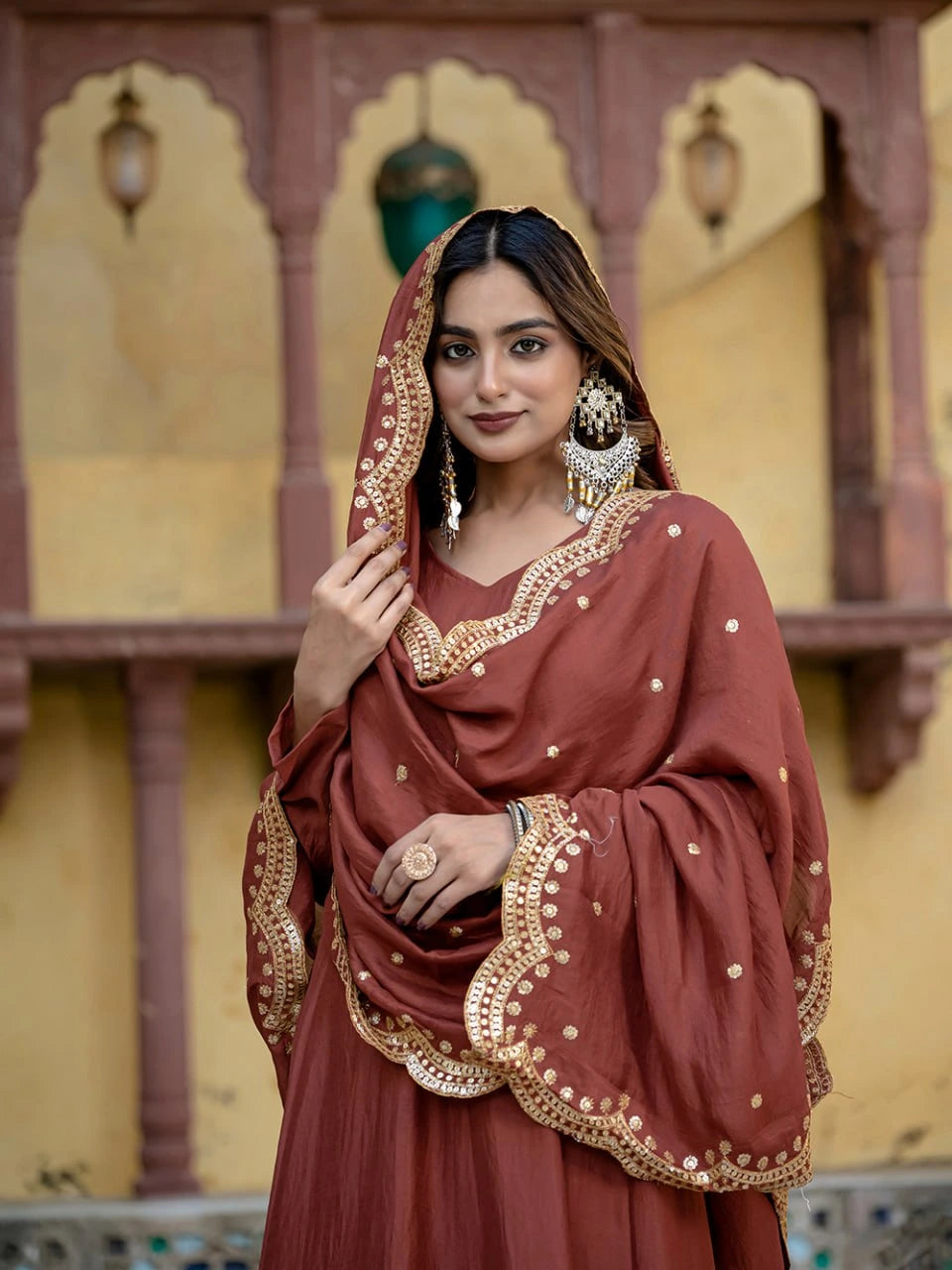 Woman in traditional maroon outfit with gold embroidery against a decorative wall.