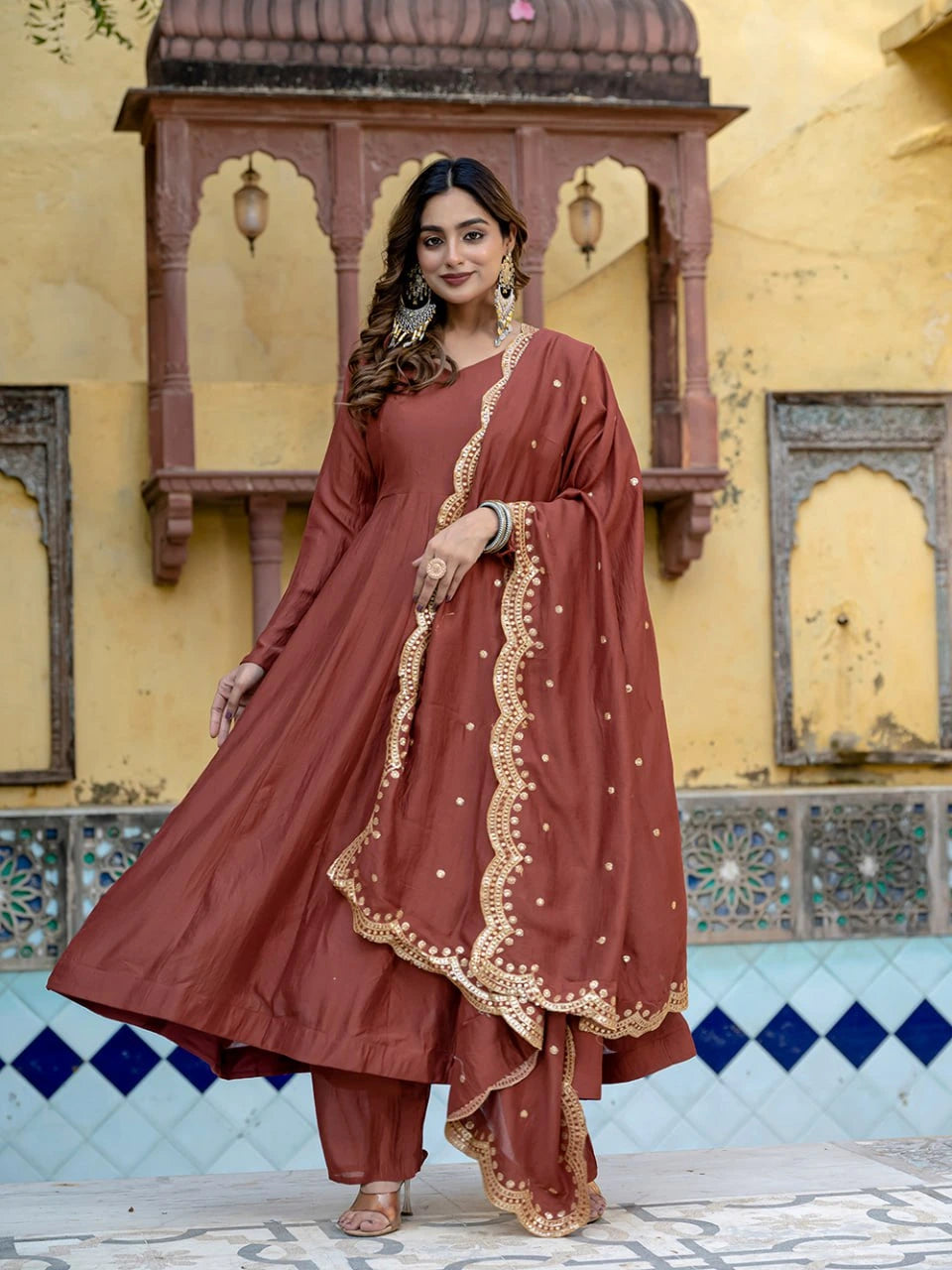 Woman in a traditional maroon outfit with gold embroidery standing in front of a decorative wall.