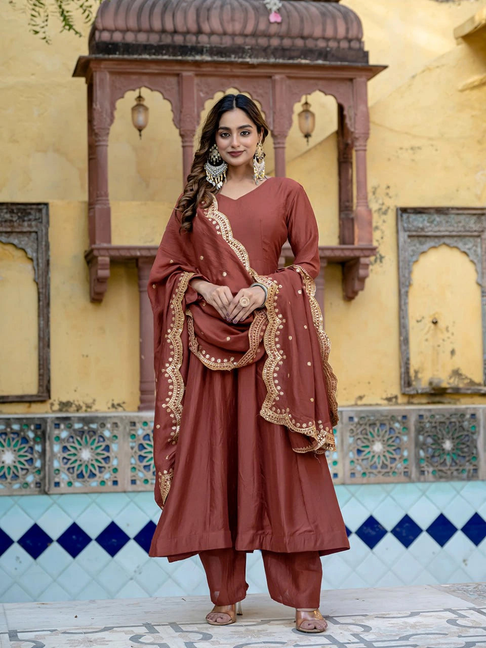 Woman in traditional brown outfit with gold details standing in front of a decorative wall.