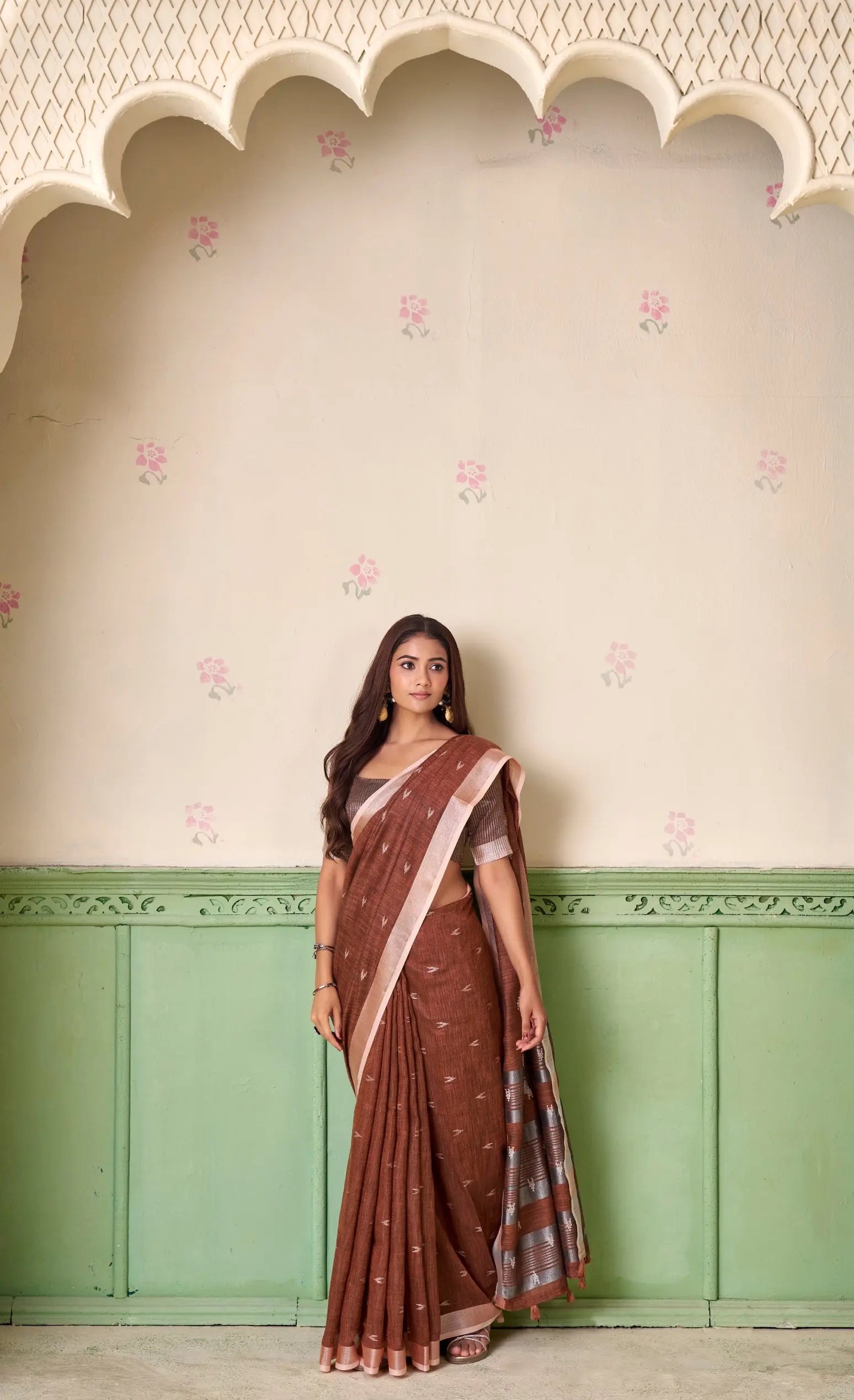 Woman wearing a brown saree with a pink border against a decorative wall.