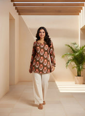 Woman wearing a patterned top and white pants standing in a well-lit room with a plant in the background.