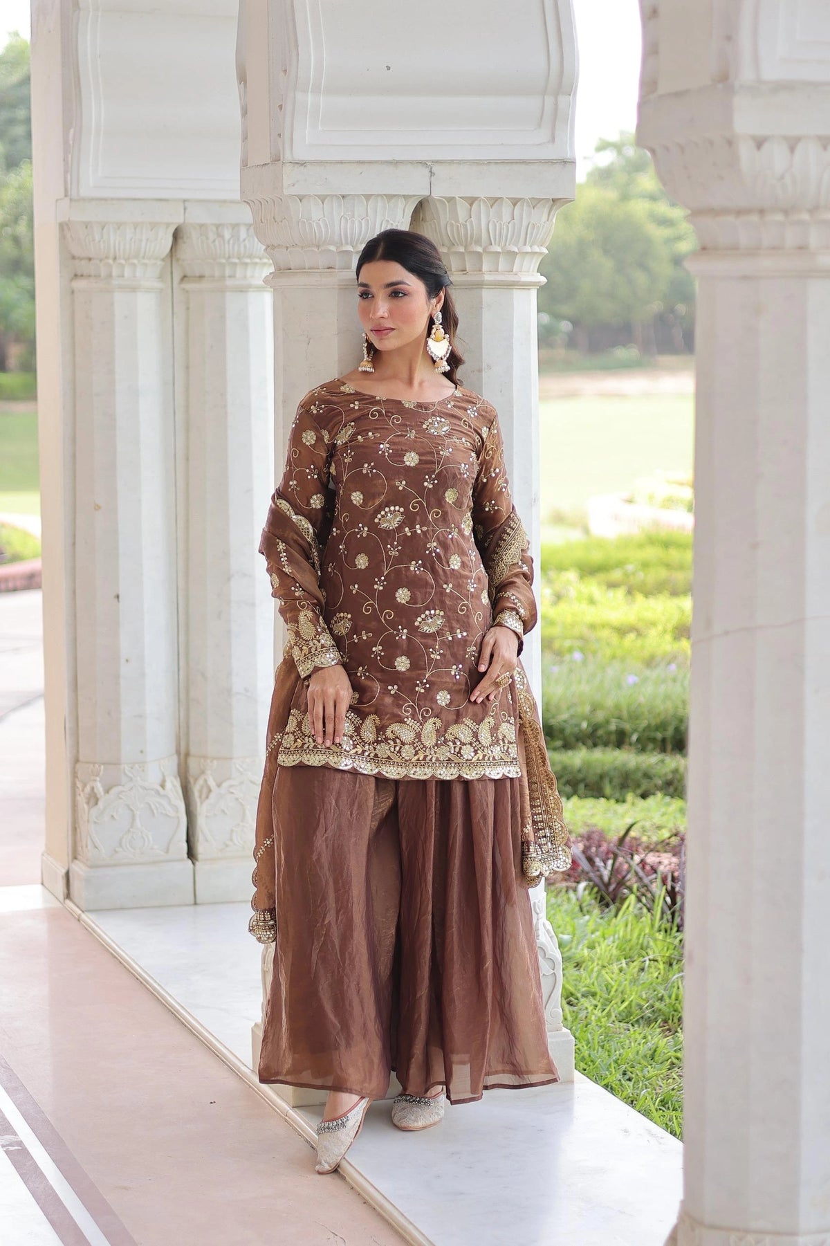 Woman in traditional brown outfit standing in front of a white architectural structure with greenery in the background