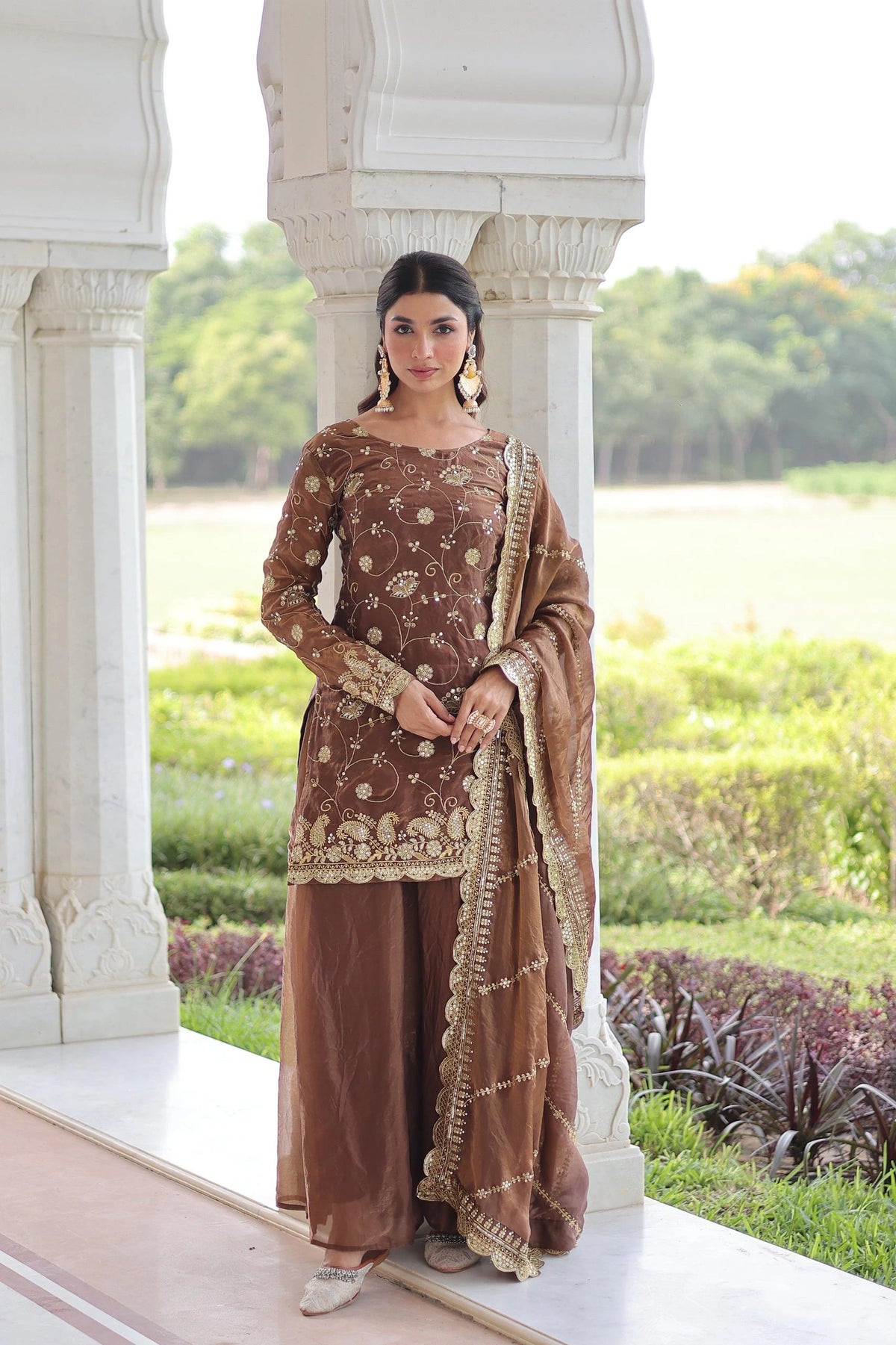 Woman in traditional brown and gold outfit standing outdoors with greenery in the background