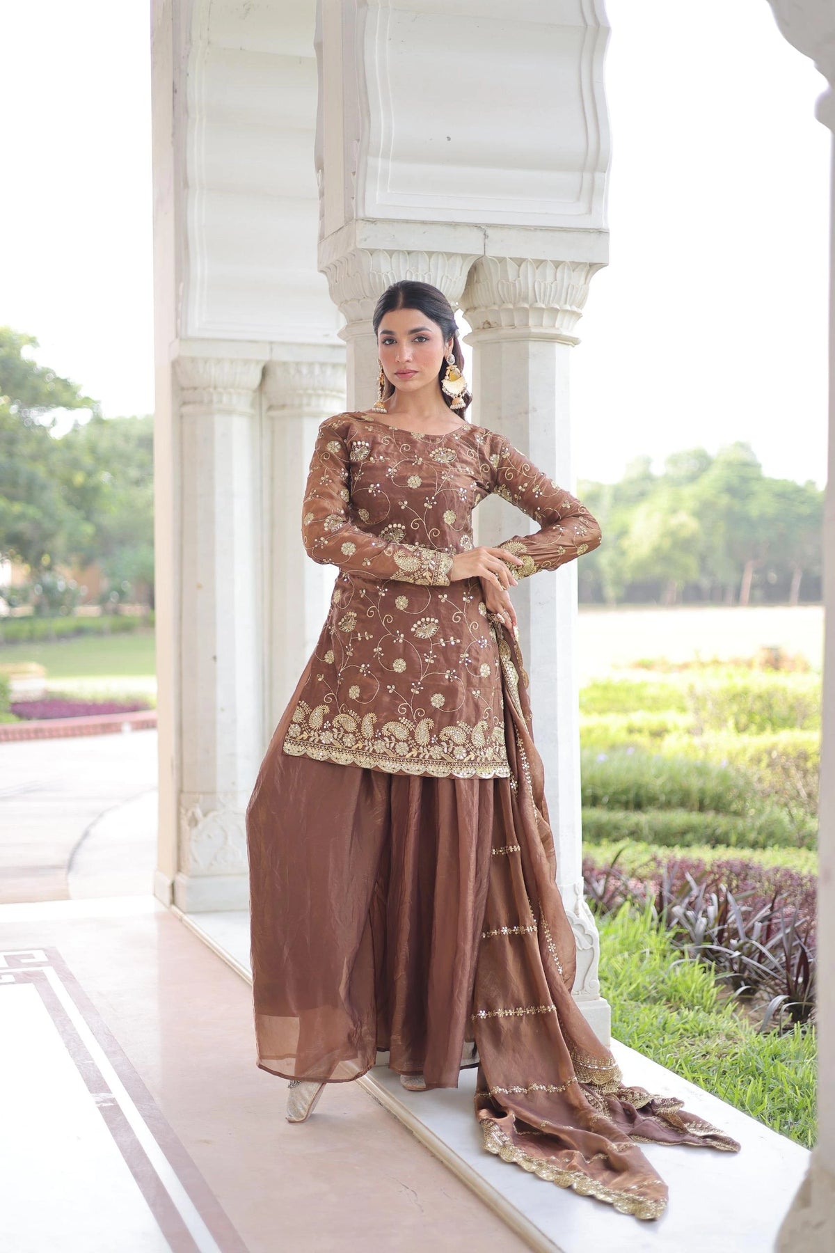Woman in a brown traditional outfit standing in front of a white architectural structure with greenery in the background.