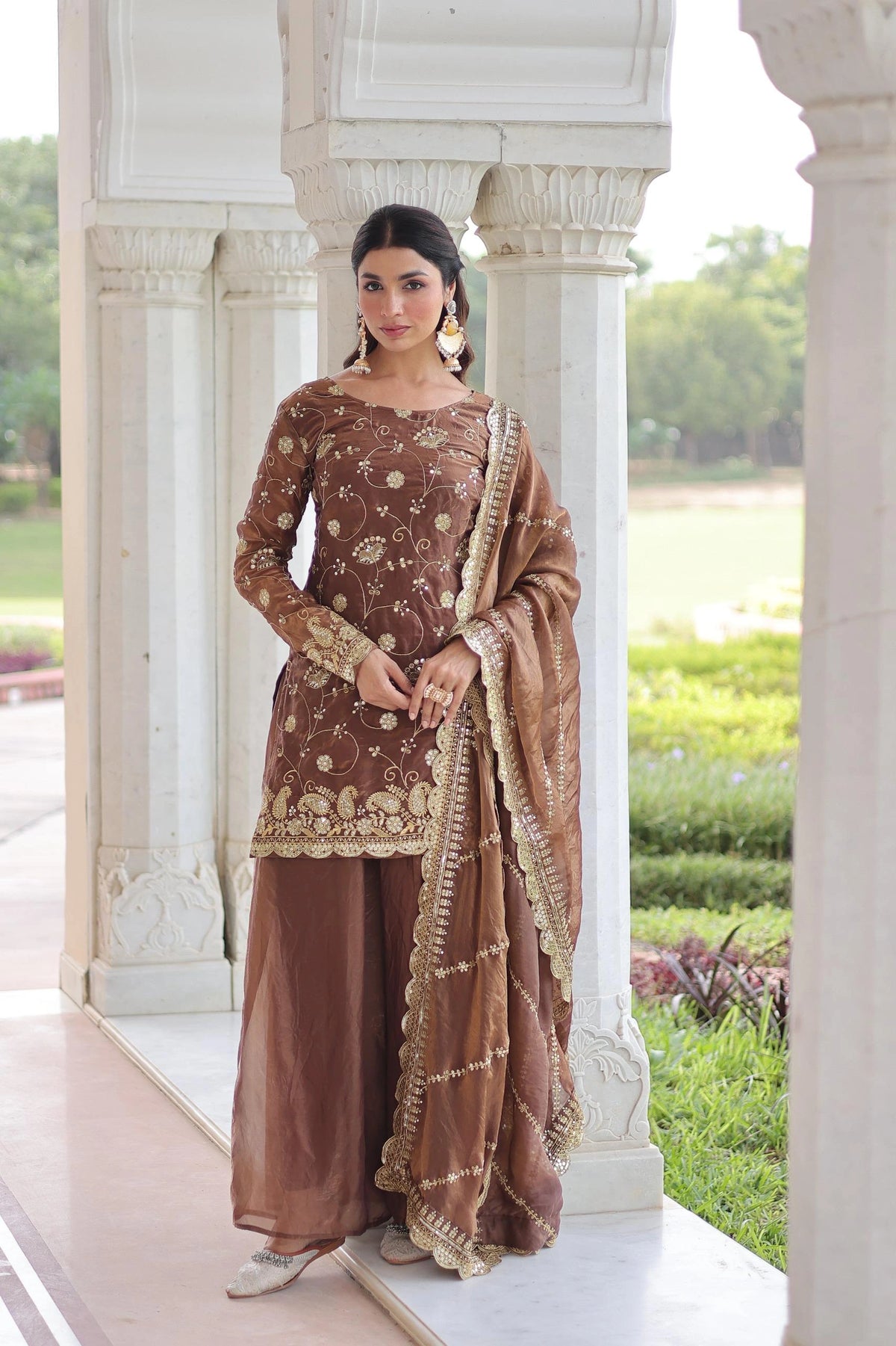 Woman in a traditional brown and gold outfit standing in front of classical architecture with greenery in the background.