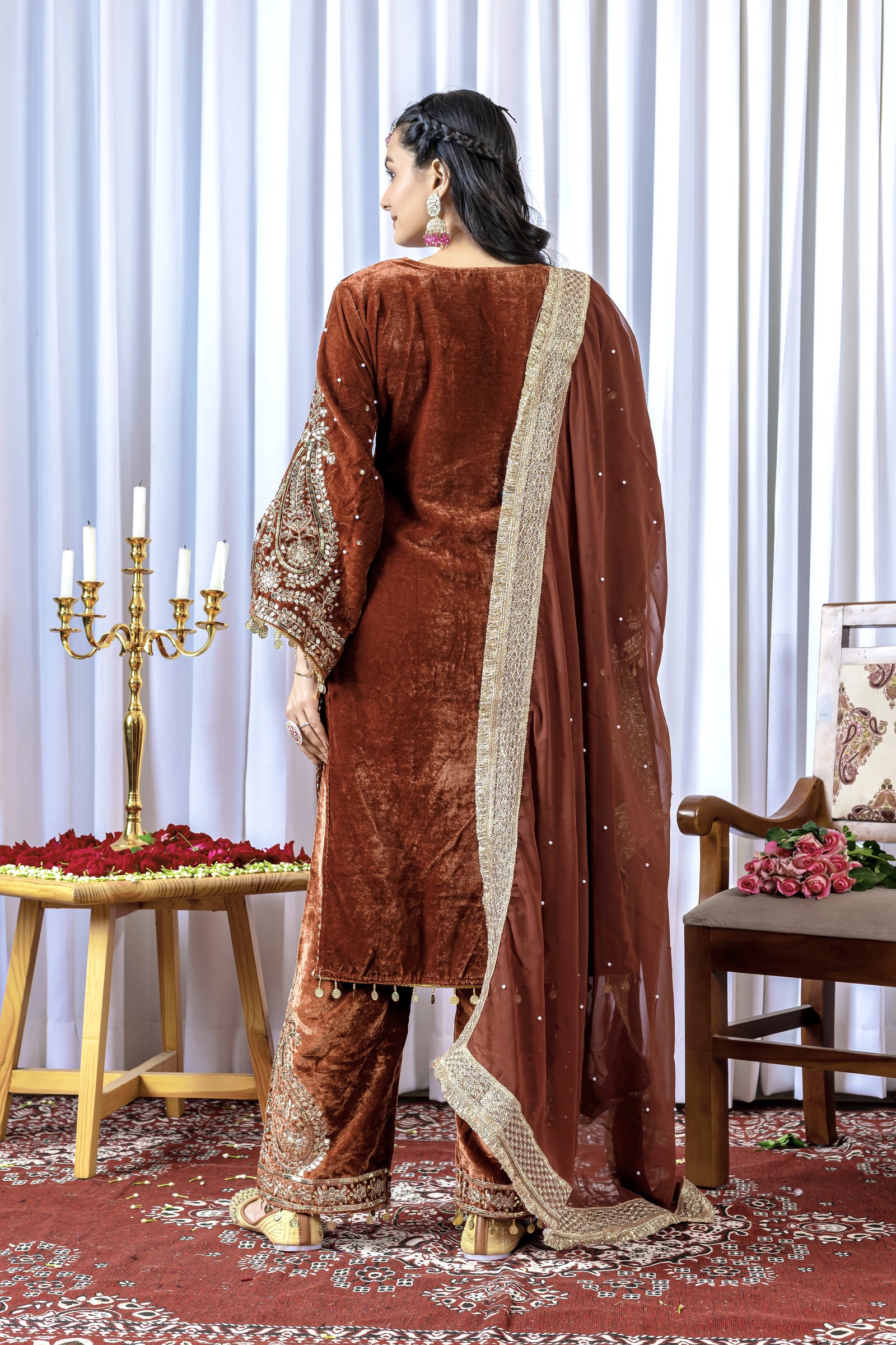 Woman in a brown traditional outfit with a matching dupatta standing in a decorated room.