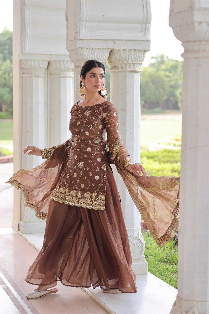 Woman in a traditional brown and gold outfit standing in front of a white architectural structure with greenery in the background.