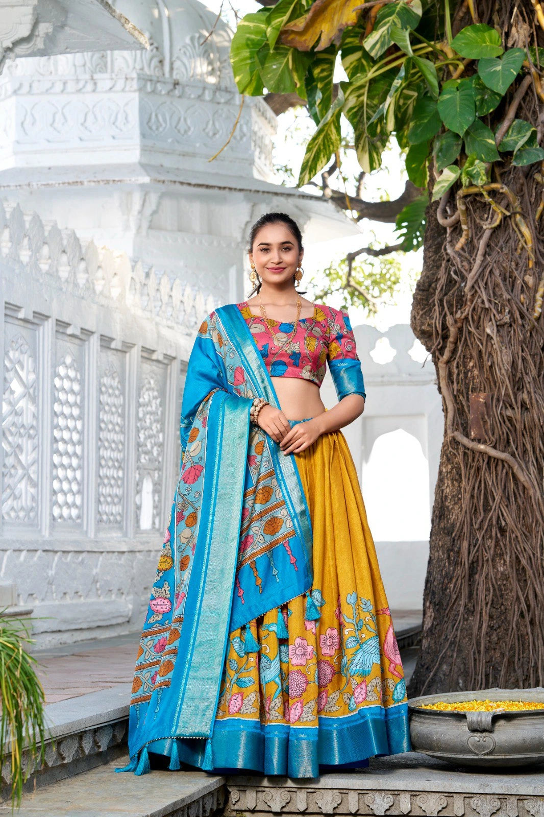 Woman wearing a vibrant mustard yellow floral lehenga with a matching printed blouse and a blue silk dupatta featuring elegant woven borders and tassel details.