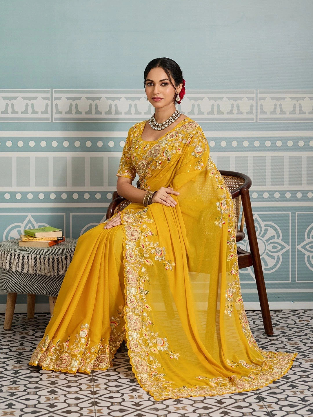 Woman in a yellow saree with floral patterns sitting on a chair against a decorative wall.
