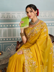 Woman in a yellow saree holding a book against a decorative wall.