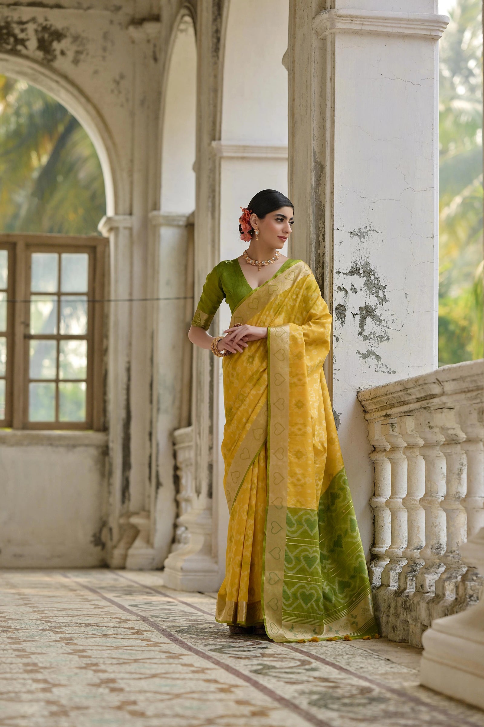 Woman in a yellow and green saree standing in an elegant indoor setting.